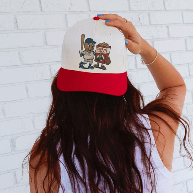 Peanut and Cracker Jack Character - PICK YOUR TEAM DTF Heat Transfer, person wearing a baseball cap with a Nut and Cracker Jack mascot design against a white brick wall background.