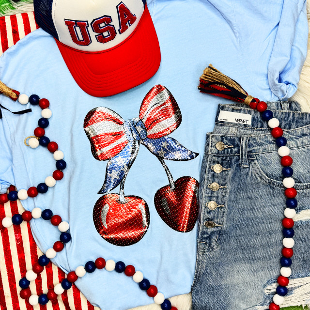 Patriotic Cherries with Bow Sequin Transfer, close-up of a light blue T-shirt with a sequin cherry graphic and bow design, paired with denim shorts, red, white, and blue bead necklaces, and a red, white, and blue USA cap, celebrating patriotic theme.