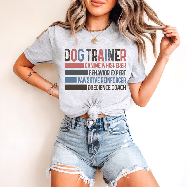 Dog Trainer Canine Whisperer Behavior Expert Pawsitive Reinforcer Obedience Coach DTF Heat Transfer, woman wearing a gray T-shirt with colorful text design for dog training and obedience coaching.