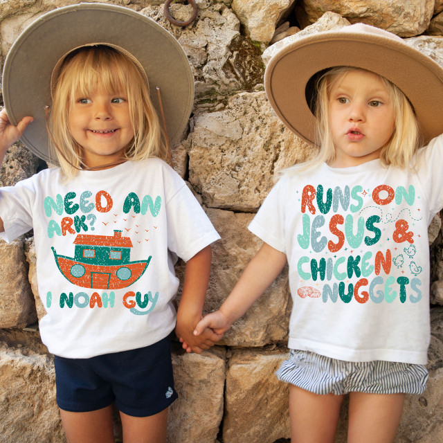 Need An Ark? I Noah Guy DTF Heat Transfer, two young girls holding hands wearing matching T-shirts with fun biblical-themed prints, one with a boat illustration and the other with playful text about Jesus and chicken nuggets, standing against a stone wall.