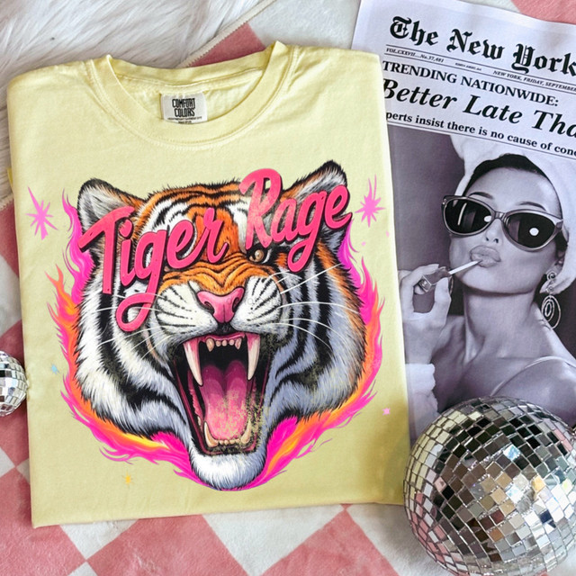 Tiger Rage DTF Heat Transfer, vibrant graphic of a roaring tiger with pink and orange flames on a yellow T-shirt, displayed with a newspaper and a decorative disco ball.