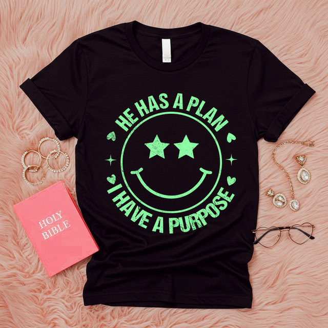 He Has A Plan I Have A Purpose GREEN DTF Heat Transfer T-shirt with smiley face design, black t-shirt laid flat on pink furry background with jewelry and a Bible