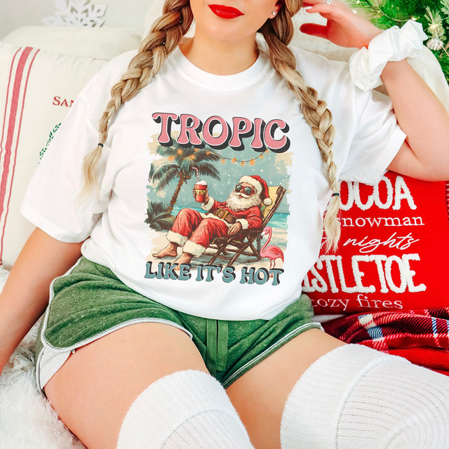 Tropic Like It's Hot DTF Heat Transfer, Christmas-themed graphic T-shirt with Santa relaxing on a beach chair holding a drink, with palm trees, a flamingo, and holiday decorations in the background.