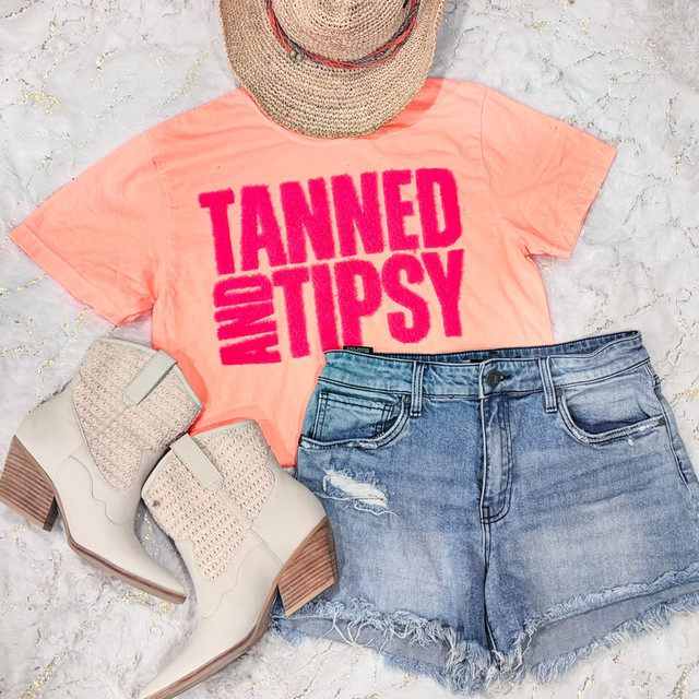 Hot Pink Tanned & Tipsy Fluffy Patch, casual summer outfit consisting of a coral pink t-shirt with bold pink text, distressed denim shorts, beige ankle boots with knitted texture, and a wide-brim straw hat with colorful bands, laid on a sandy surface.