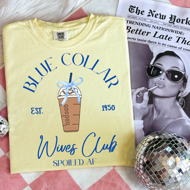 Blue Collar Wives Club DTF Heat Transfer, yellow T-shirt with blue text and an ice coffee graphic, laid flat with a newspaper and decorative disco ball.
