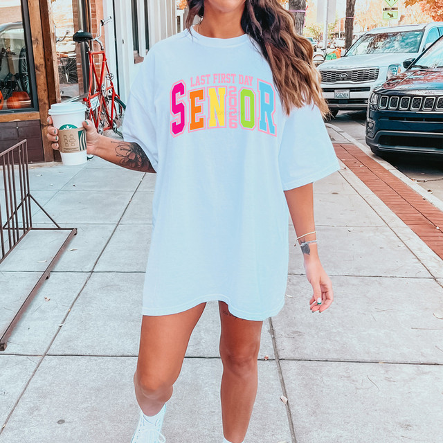 Rainbow Block Last First Day Senior 2026 DTF Heat Transfer, colorful graphic t-shirt worn by a woman holding a coffee cup on a city sidewalk.