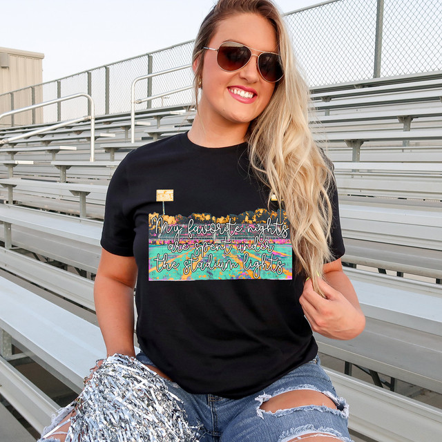 My Favorite Nights Are Spent Under the Stadium Lights DTF Heat Transfer, a woman wearing a black T-shirt with a colorful stadium scene and text, sitting in stadium bleachers with sunglasses and holding pom-poms.