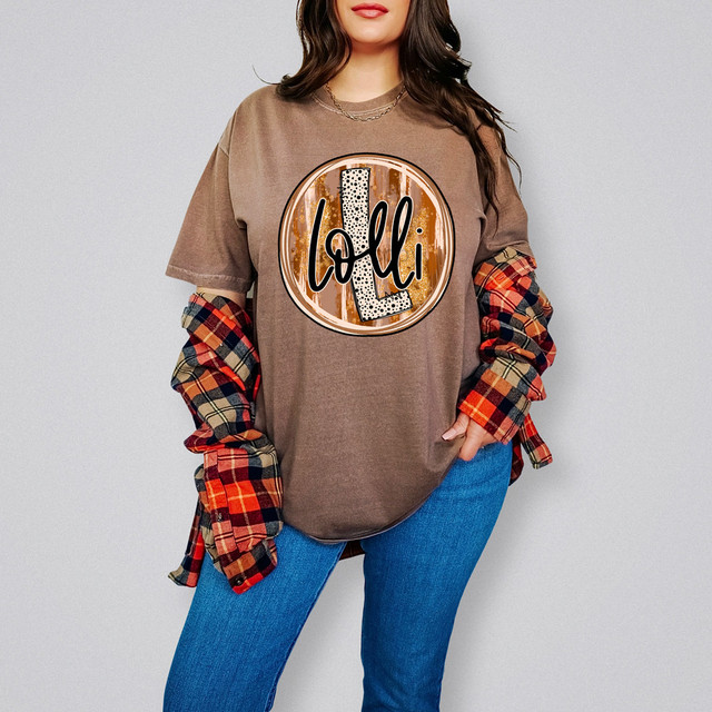 Lolli Fall Brushstrokes DTF Heat Transfer, a digitally printed heat transfer design with fall-themed brushstrokes and the word "Lolli" in a playful, dotted font on a brown T-shirt worn by a woman.