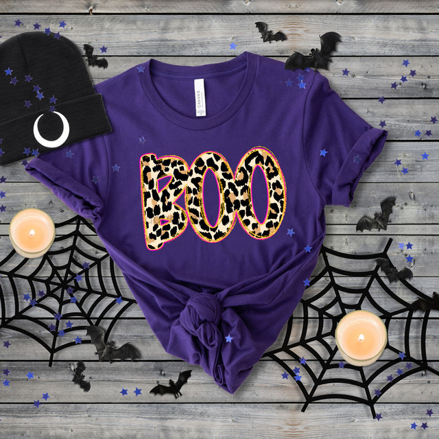 Leopard Boo DTF Heat Transfer, purple T-shirt with leopard print 'BOO' design on a wooden surface decorated with Halloween themed items.