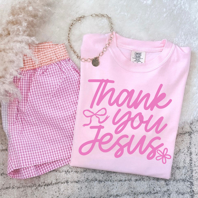 Thank You Jesus PINK DTF Heat Transfer, pink T-shirt with thank you Jesus text in pink and flower emblem, next to pink and red gingham shorts and a silver chain necklace on a light furry surface.