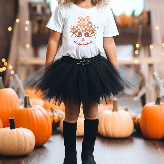 Whimsical Scarecrow DTF Heat Transfer, fun Halloween-themed pumpkin costume with girl wearing a white t-shirt featuring a scarecrow face design with pumpkin hat and black tulle skirt surrounded by pumpkins.