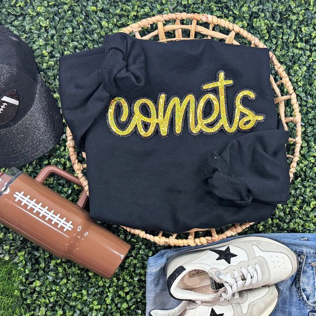 Comets Mascot Gold Sequins Patch, a black hoodie with vibrant gold sequin embroidery spelling 'comets' displayed on a textured green surface with other athletic accessories.
