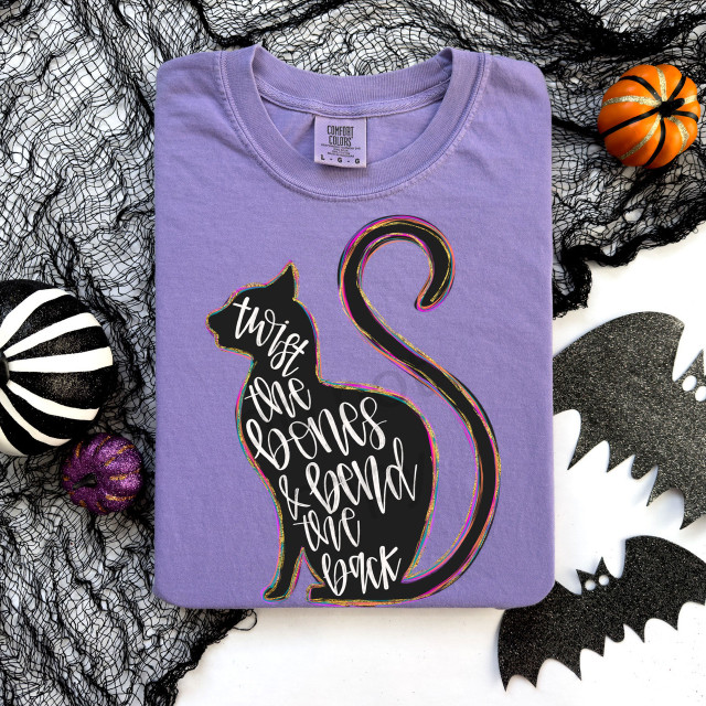 Twist The Bones & Bend The Back DTF Heat Transfer, Halloween-themed design on a folded lavender t-shirt with black, orange, and glittery Halloween decorations around it.