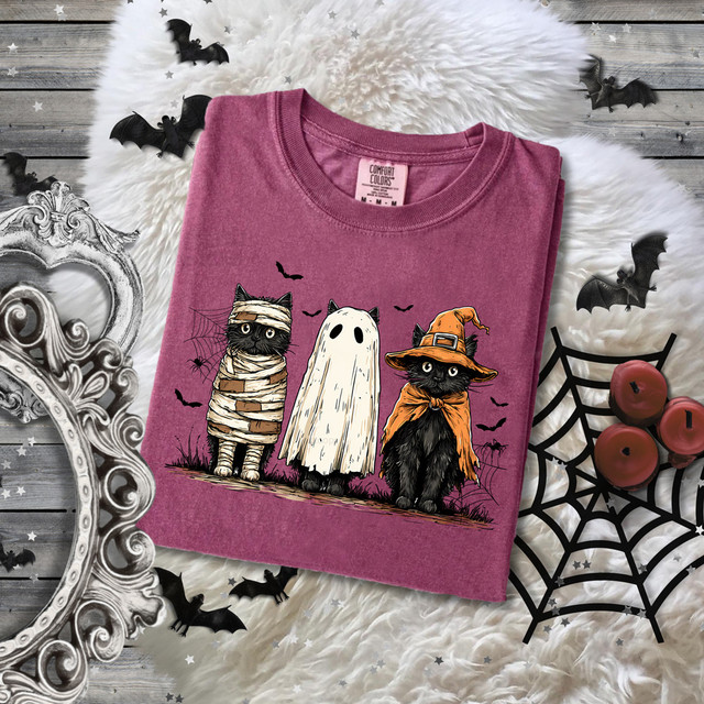 Black Cat Halloween Trio DTF Heat Transfer, featuring three cute black cats dressed as a mummy, ghost, and witch on a pink t-shirt with Halloween decorations around.