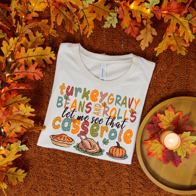 Thanksgiving Casserole DTF Heat Transfer, seen on a white t-shirt with colorful autumn-themed text and illustrations of a casserole dish, turkey, and pumpkin, placed on a textured orange blanket with fall leaves and a candle.