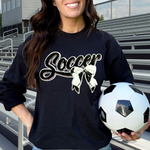 Soccer With Bow Faux Patch DTF Heat Transfer, close-up of a black sweatshirt featuring a patch of the word 'Soccer' with a bow design on it, worn by a woman holding a soccer ball in an outdoor stadium.