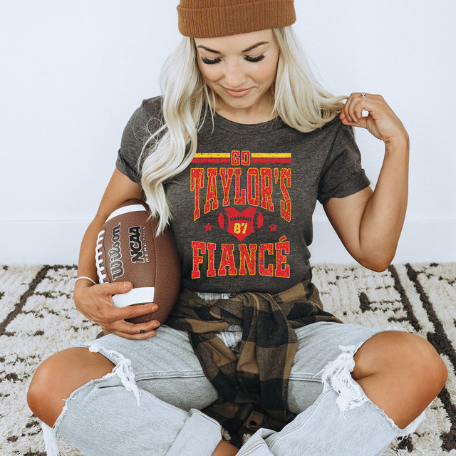 Go Taylor's Fiance DTF Heat Transfer, woman wearing a dark t-shirt with red and yellow design, sitting on the carpet, holding a football.