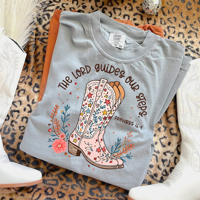 The Lord Guides Our Steps DTF Heat Transfer, a graphic design on a folded gray T-shirt featuring a cowboy boot with floral and star patterns, surrounded by decorative floral elements and the Bible verse Proverbs 16:9.
