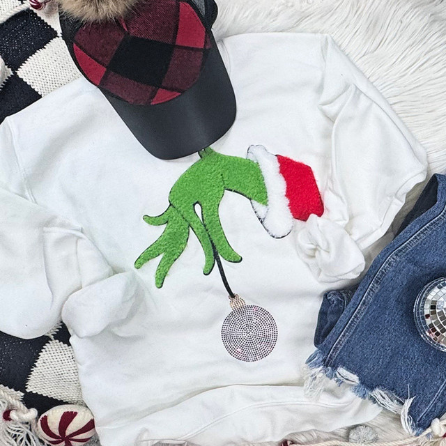 Fluffy Hand Holding an Ornament Patch, Christmas themed fluffy green hand with a red and white Santa hat ornament patch on a white sweatshirt, holiday fashion accessory.
