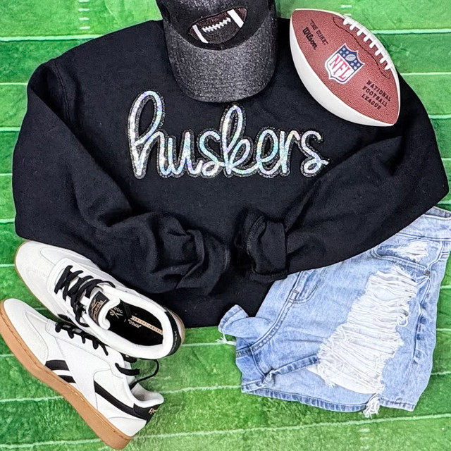 Huskers Cursive Sequin Patch, black sweatshirt with 'huskers' written in colorful sequins, paired with ripped denim shorts, white sneakers, and a baseball cap, sports-themed outfit on a green background.