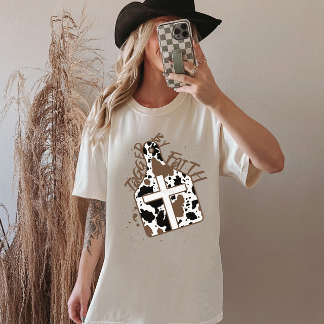 Tagged By Faith DTF Heat Transfer, woman wearing a T-shirt with a cow print cross design and cow print tag icon, styled with a black hat and taking a mirror selfie.
