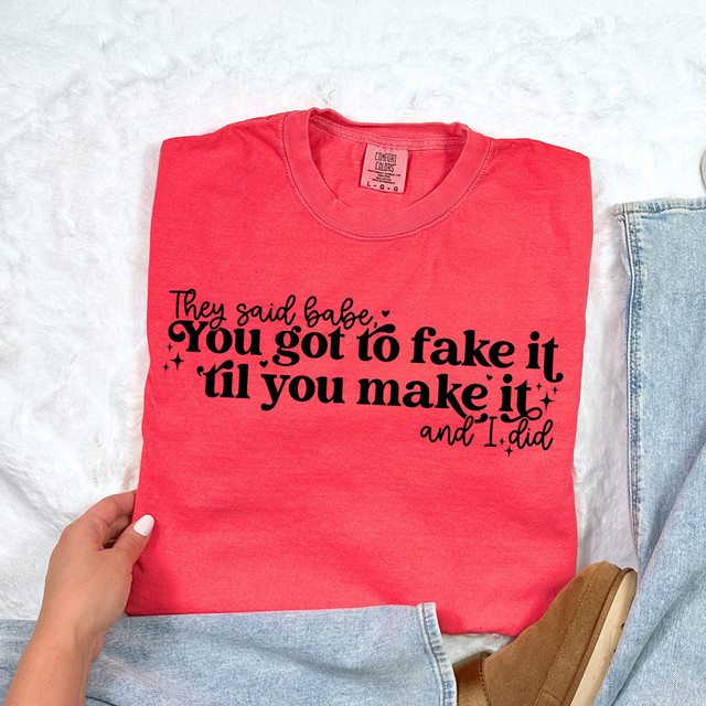 Fake It Til You Make It BLACK DTF Heat Transfer, image of a red t-shirt with black text and decorative stars, ready for application onto fabric.