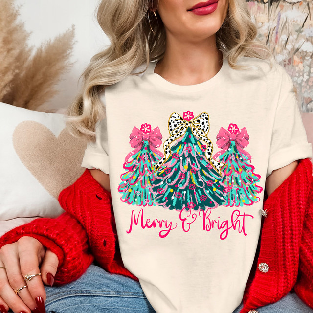 Merry & Bright Preppy Trees DTF Heat Transfer, colorful Christmas trees featuring bows and floral details with the text 'Merry & Bright' in pink, on a woman's casual outfit.