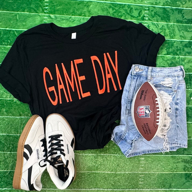 FLAWED Large Game Day PUFF Embroidered Patch - Various Colors, casual outfit featuring a black T-shirt with 'GAME DAY' printed in bold red letters, distressed denim shorts with NFL football patch, and white Reebok sneakers, laid out on a green wooden surface.