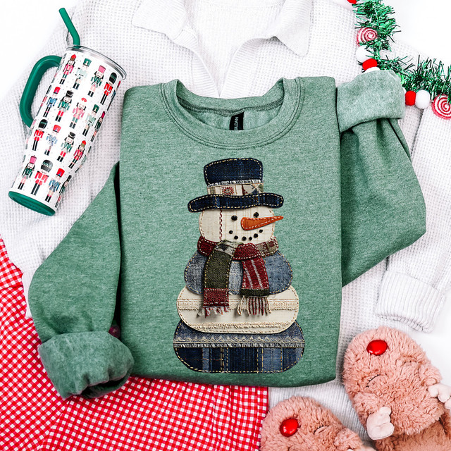 Patchwork Snowman DTF Heat Transfer, decorative snowman made of various fabric patches with a top hat, scarf, and carrot nose on a green sweatshirt surrounded by holiday decorations.