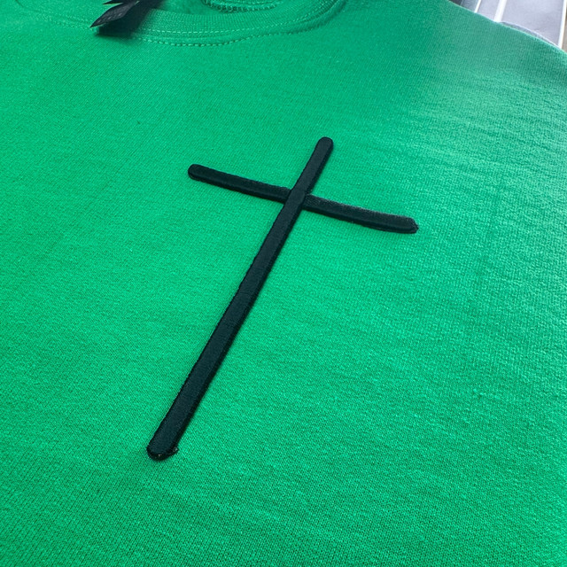 Cross Oversized Pocket Embroidered Patch, close-up of a green sweatshirt with a black cross patch embroidered on the front.