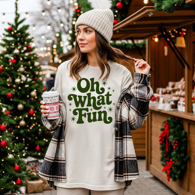 Retro Oh What Fun GREEN DTF Heat Transfer, a woman wearing a Christmas themed sweatshirt with the text 'Oh what Fun' in green surrounded by snowflakes, holding a festive coffee cup against a holiday market backdrop.