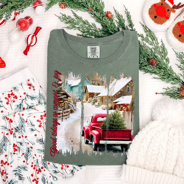 Good Tidings Of Comfort & Joy DTF Heat Transfer, a Christmas-themed t-shirt with a winter scene of a snowy village and a red truck carrying a Christmas tree, surrounded by holiday decorations.