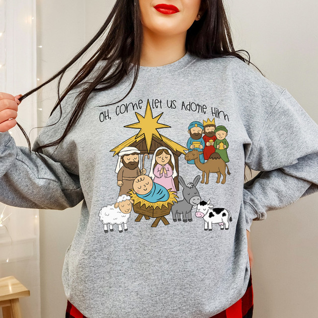 Oh Come Let Us Adore Him Nativity Illustration DTF Heat Transfer T-shirt worn by a woman with long dark hair and red lipstick, featuring a cartoon nativity scene with baby Jesus, Mary, Joseph, the Wise Men, shepherds, angels, animals, and a star, in a colorful design.