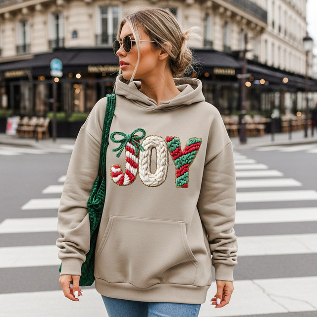 Faux Knit Joy Patterned DTF Heat Transfer, a woman's beige hoodie with colorful embroidered holiday-themed letters spelling 'JOY' with festive yarn and ribbon details.
