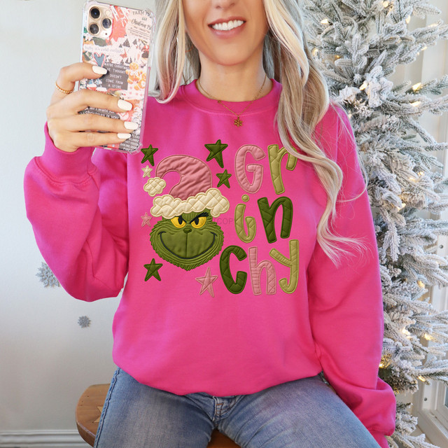 Faux Fabric Green Guy DTF Heat Transfer, cheerful woman wearing pink Christmas sweatshirt with Grinch face design and holiday-themed embroidery.