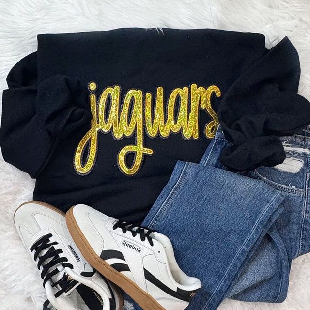 Jaguars Gold Sequin Patch, black sweatshirt with yellow glittered text and sequined jaguar patch, paired with blue jeans and white Reebok sneakers with black accents.