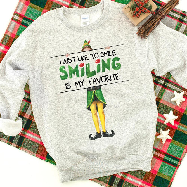 I Just Like To Smile Smiling Is My Favorite Screen Print Heat Transfer sweatshirt, featuring a festive design of a cheerful elf holding a sign with the quote, displayed on a plaid background with holiday decorations.