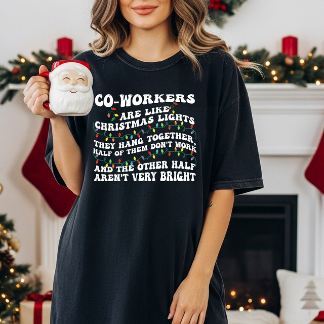 Coworkers Are Like Christmas Lights DTF Heat Transfer, a woman wearing a black T-shirt with Christmas lights and humorous text, holding a Santa mug, in a festive holiday setting.