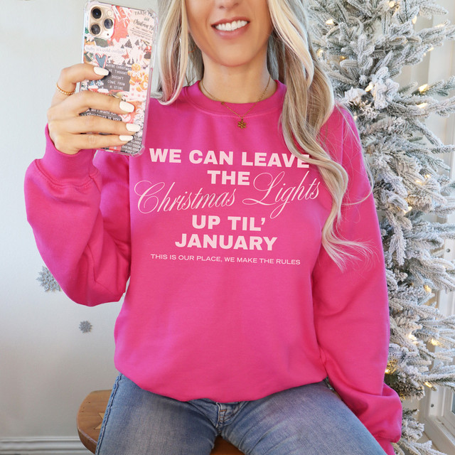 We Can Leave the Christmas Lights Up Til’ January PINK DTF Heat Transfer, a woman wearing a pink sweatshirt with white and pink lettering, holding a decorated phone with a Christmas tree in the background.