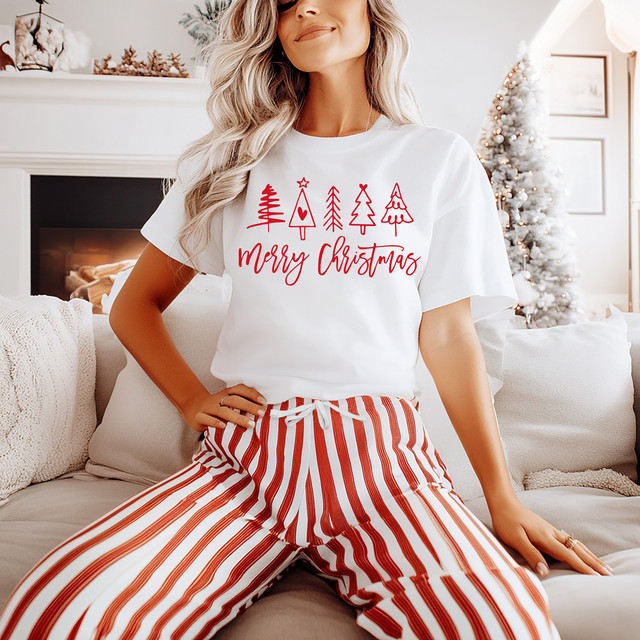 Merry Christmas Minimalist Pattern Trees RED DTF Heat Transfer, a woman wearing a white Christmas t-shirt with red tree patterns and red text, in a cozy living room with a Christmas tree in the background.