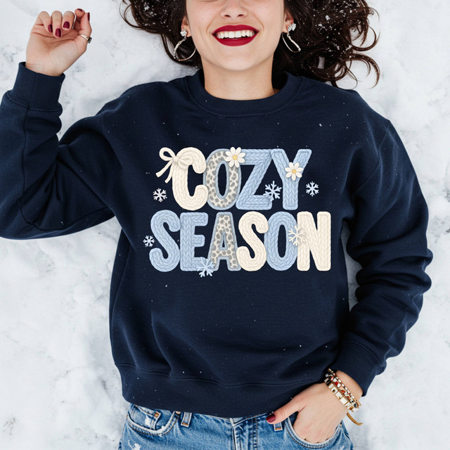 Faux Yarn Cozy Season DTF Heat Transfer, a woman wearing a navy blue sweatshirt with a design that says 'Cozy Season' in pastel colors and winter-themed accents like snowflakes and flowers.