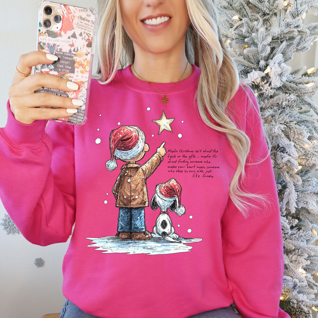 Christmas Scene Black/White Dog DTF Heat Transfer, a woman wearing a pink sweatshirt with a cartoon of a girl and a dog in Santa hats, reaching for a star on a Christmas tree.