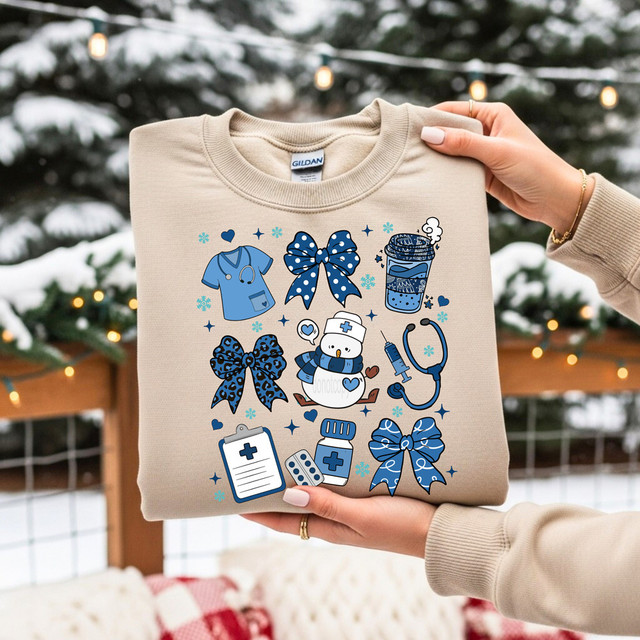 Winter Nurse Collage DTF Heat Transfer, a beige sweatshirt with medical-themed cartoon illustrations in blue and white, including a nurse snowman, medical bows, stethoscope, syringe, medicine bottle, clipboard, and pills, on a winter outdoor background.