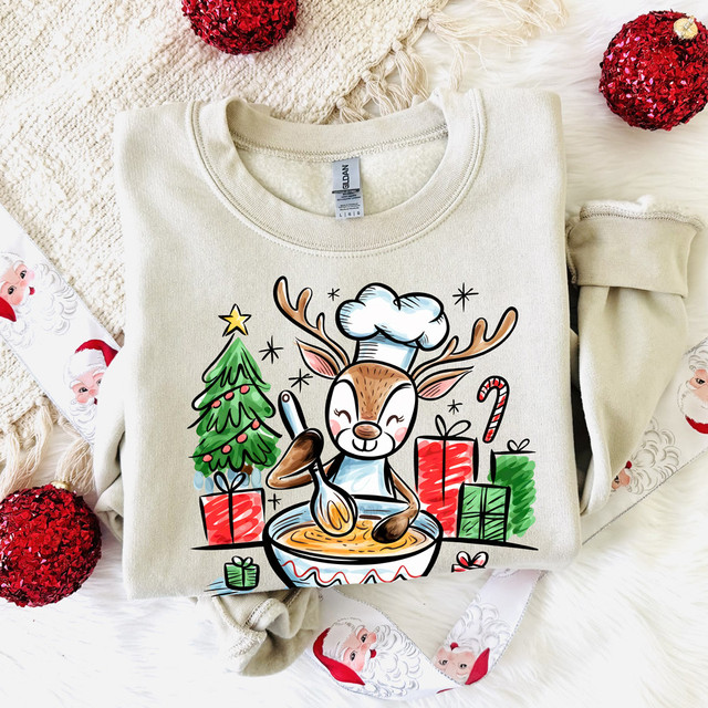 Reindeer Baking DTF Heat Transfer, illustrated reindeer chef with a Christmas tree and presents on a sweatshirt, perfect for holiday crafts and Christmas themed clothing.