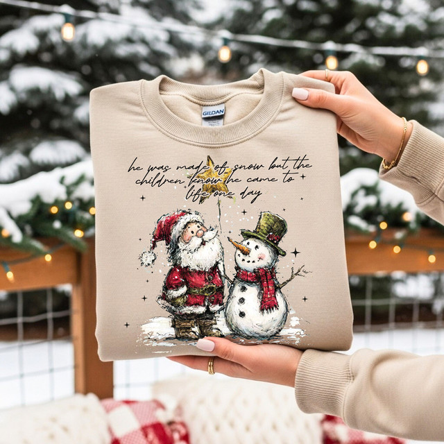 He Was Made Of Snow But The Children Know He Came To Life One Day, Christmas-themed heat transfer design on beige sweatshirt, featuring Santa Claus and a snowman with festive text.