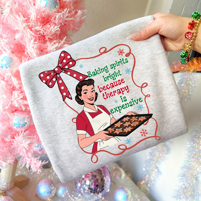 Baking Spirits Bright Because Therapy Is Expensive DTF Heat Transfer, a humorous holiday-themed graphic on a gray t-shirt featuring a woman holding a tray of cookies, with festive decorations and a pink Christmas tree in the background.