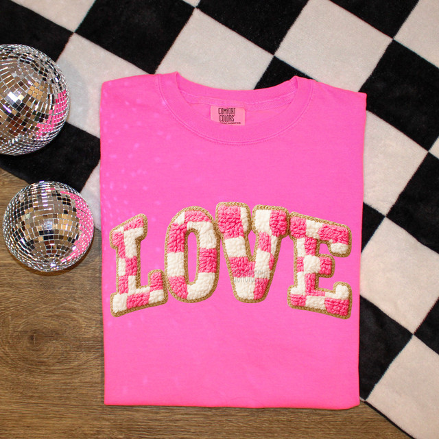 Faux Knit Pink Checkered Love DTF Heat Transfer, pink t-shirt with embroidered love design, placed on a wooden surface with checkered backdrop and disco balls, showcasing a trendy fashion accessory.