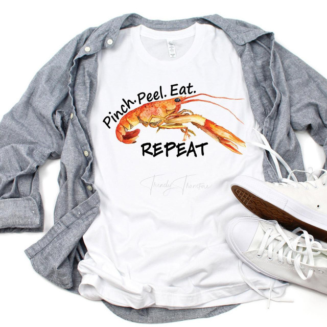It's arm Day T-shirt, featuring a shrimp graphic with the text 'Pinch. Peel. Eat. REPEAT' in a casual style, laid out on a flat surface with sneakers and a gray shirt.