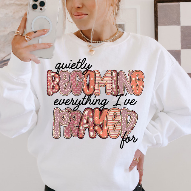 Quietly Becoming Everything I've Prayed For DTF Heat Transfer, a woman wearing a white sweatshirt with colorful, patterned text design.