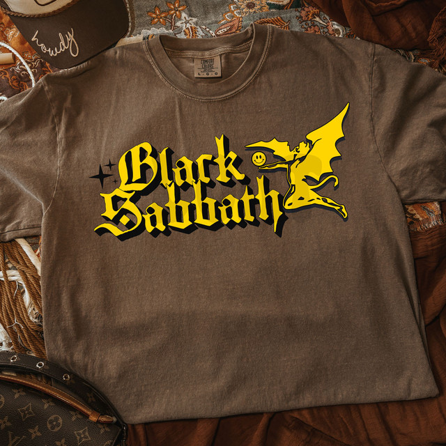 Sixties Band Tee DTF Heat Transfer, vintage brown T-shirt with 'Black Sabbath' logo and yellow graphic design of a demon with wings and a smiley face, on a textured fabric background.